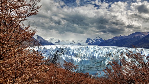 Blue glacier in the middle of a mountainous landscape with dramatic clouds and autumn leaves, The