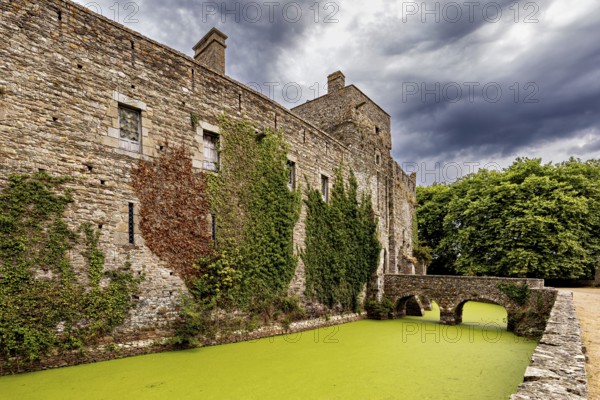 Side view of a castle with a bridge over a moss-covered moat under a cloudy sky, Pirou Castle in