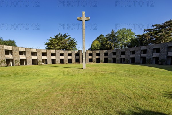 A stone cross sits enthroned in the centre of a green, circular courtyard surrounded by a concrete