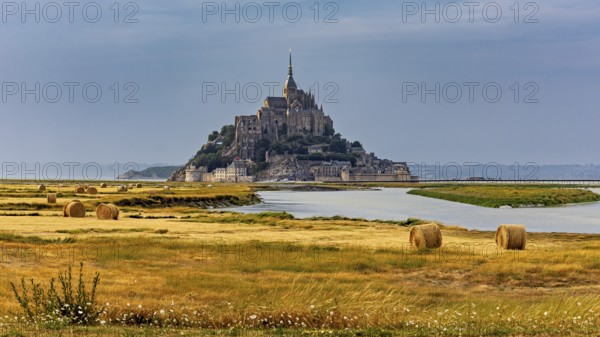 A castle rises in the middle of fields and a river, surrounded by bales of straw, Mont Saint Michel