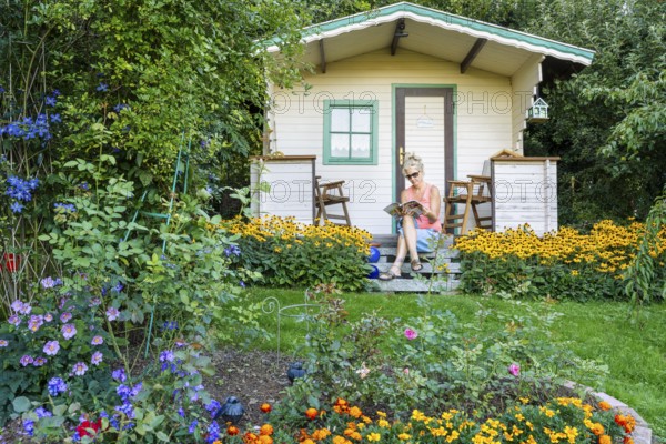 A woman sits relaxed on the small veranda of a garden shed and reads. Blooming summer flowers in