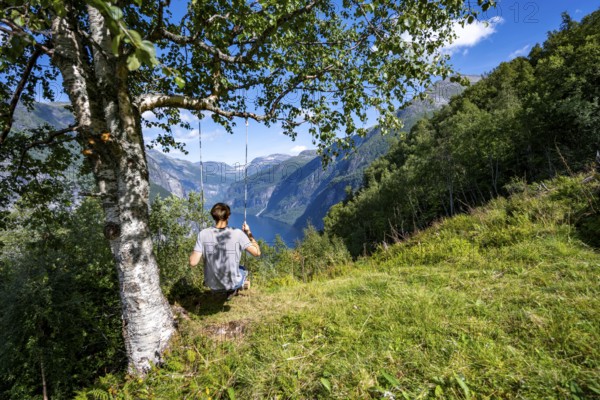 Young man swinging on a tree, view of idyllic fjord landscape of the Geirangerfjord at Blomberg