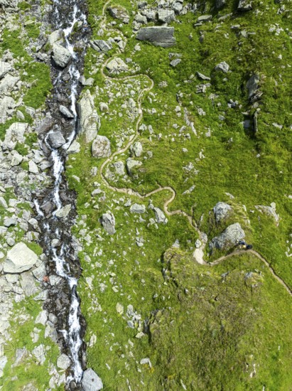 Hiking trail winds through mountain meadows interspersed with stones next to a mountain stream,