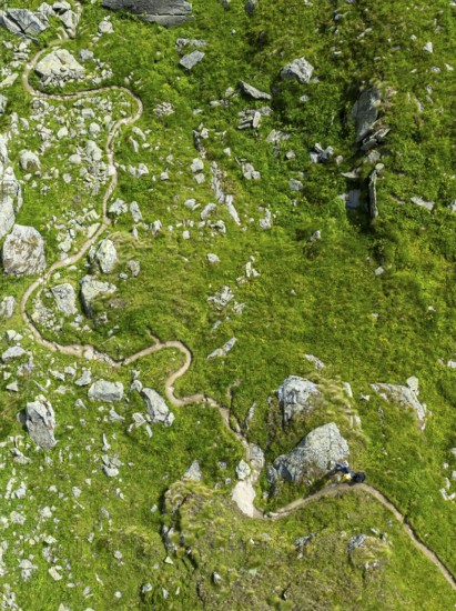 Hiking trail winds through mountain meadows interspersed with stones, from above, aerial view,