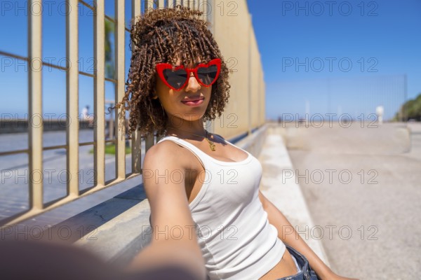 Fashionable young woman with curly hair enjoying a sunny day, taking a selfie by a stylish metal