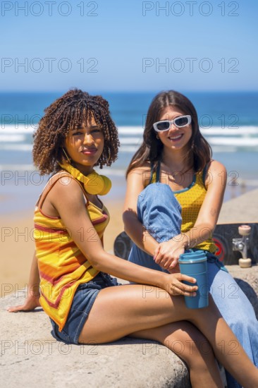 Two young women relaxing on beach wall, enjoying ocean view with skateboard and reusable coffee