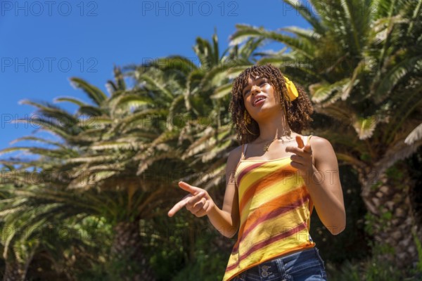 Carefree young woman with headphones enjoying music and pointing while standing under palm trees on