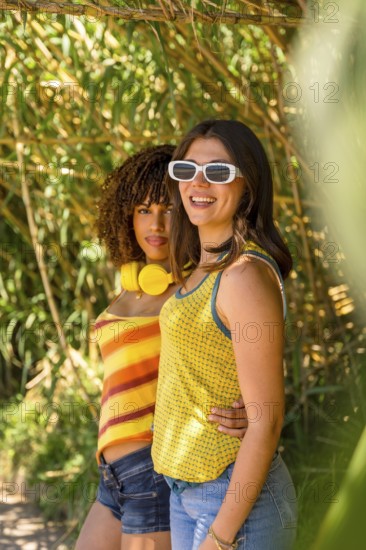 Two young women with headphones and sunglasses are embracing and smiling in a bamboo forest,