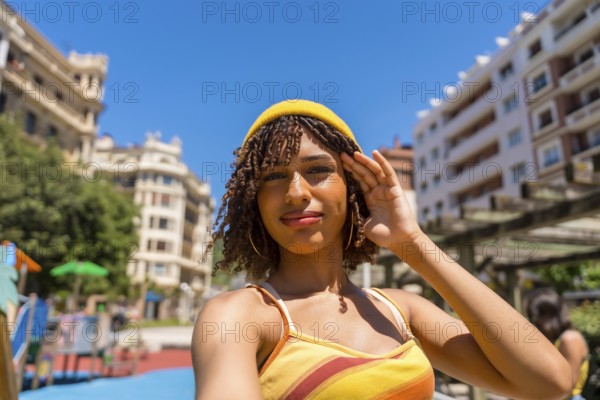 Stylish young woman with curly hair and yellow beanie taking a selfie in a sunny city park,