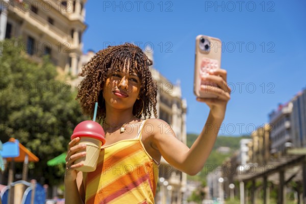 Stylish young woman captures a selfie while holding a refreshing beverage, enjoying a sunny day in