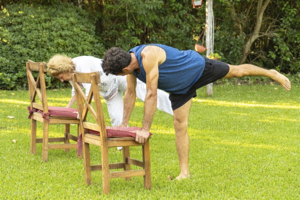 Senior man and woman practicing yoga in garden, doing balancing pose using chairs for support,