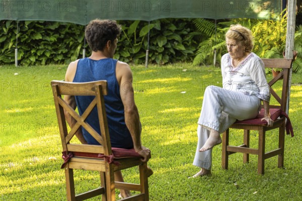 Senior woman and her physiotherapist are sitting on chairs in a garden, practicing rehabilitation