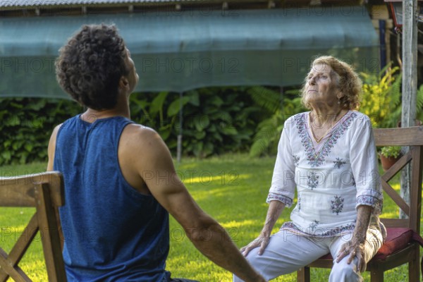 Senior woman and her physiotherapist are sitting on chairs in a garden, practicing breathing