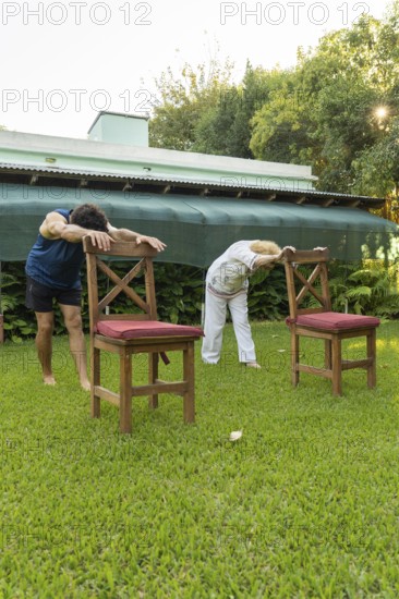 Senior woman and her physiotherapist are bending forward, holding onto chairs in a garden,