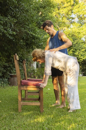 Physiotherapist is assisting a senior woman performing bending exercises with the help of a chair