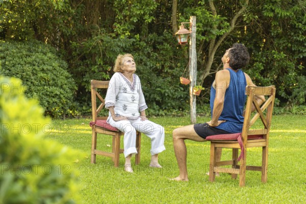 Senior woman and her physiotherapist are sitting on chairs in a garden, doing neck stretching