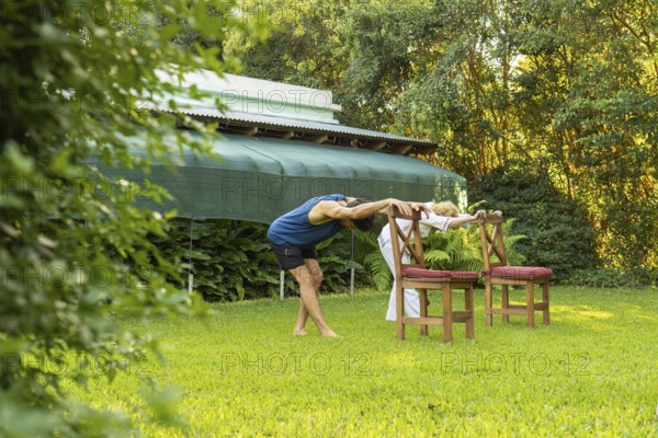 Senior couple practicing stretching exercises in their garden, using chairs as support, promoting