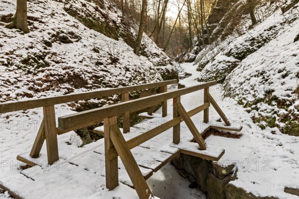 Narrow wooden bridge over a frozen stream in a snowy forest gorge at sunset, The Dragon Gorge near