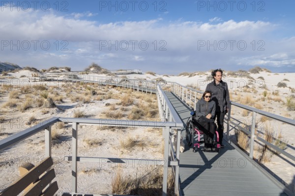 Man and handicapped woman in wheelchair stroll down the Interdune Boardwalk with their ped dog at