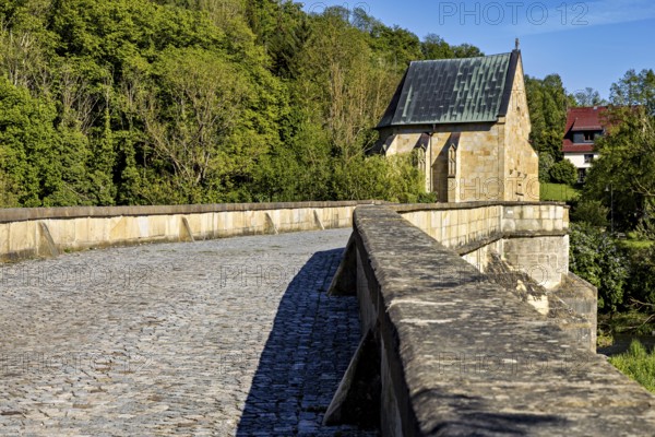 Close-up of a cobblestone road on a stone bridge leading to a chapel, surrounded by forest, The