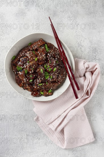 Crispy beef, with green onions and sesame seeds, in a bowl with chopsticks, on a textured surface,