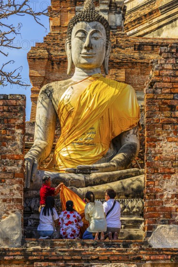 A family decorates a stone Buddha statue with yellow cloth, historic Buddhist temple complex,