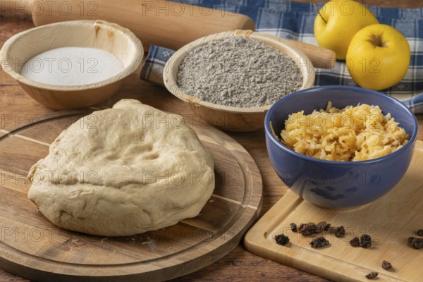 Dough prepared next to poppy seeds, grated apple and raisins on a wooden table