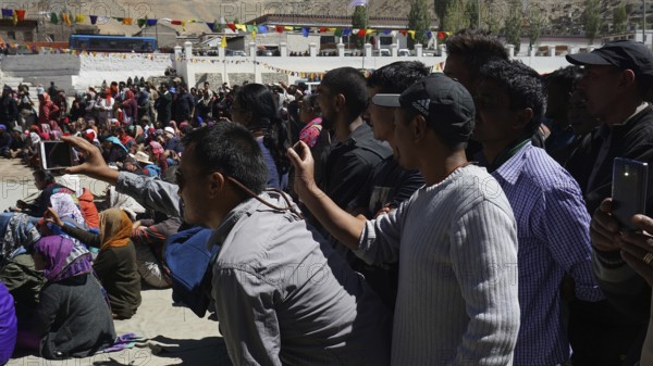 A crowd films an outdoor festival with smartphones, colorful details in the background, Leh,