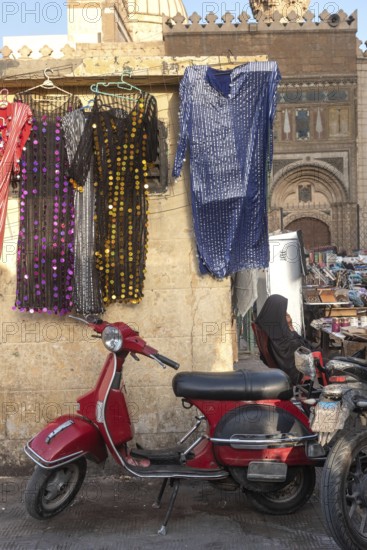 A cool looking red scooter parked amongst a busy street market selling colourful women's dresses