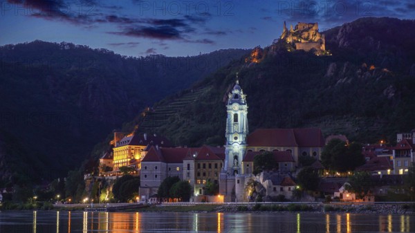 Illuminated church and castle in Dürnstein in front of a hill at dusk, reflected in the river,