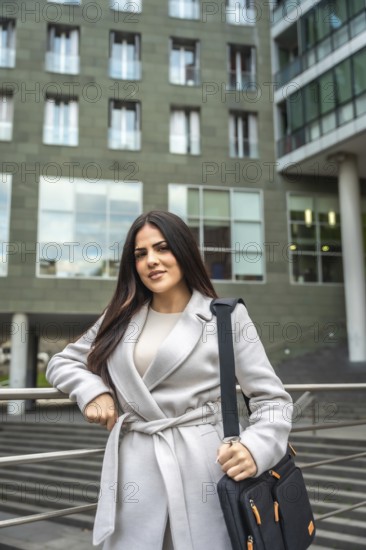 Confident businesswoman wearing a coat and carrying a shoulder bag, standing outdoors in an urban