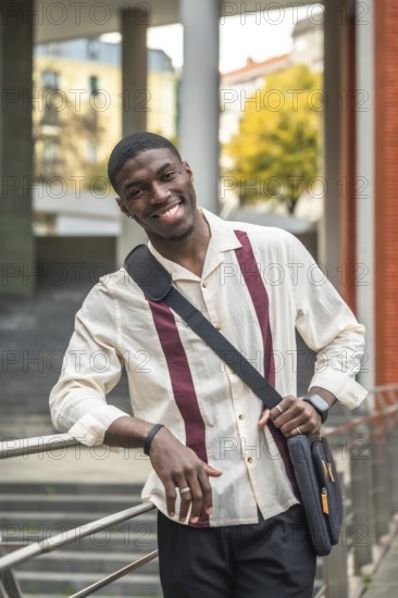 Smiling young black student on a university campus, leaning on a railing with a shoulder bag,