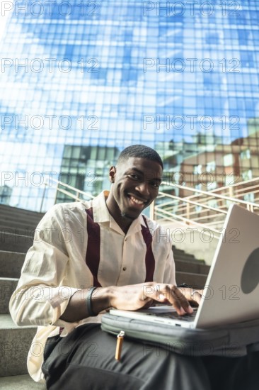 Young black man smiling while typing on a laptop, sitting on urban stairs with a modern glass