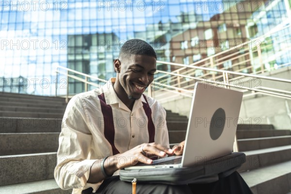 Young black man sitting on concrete stairs in a city, smiling and typing on a laptop, portraying