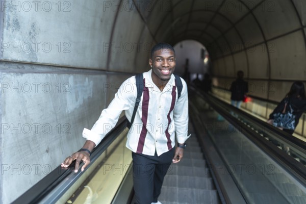 Young black man with backpack smiling at camera while holding handrail on an escalator ascending