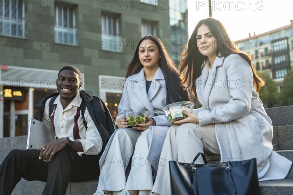 Diverse colleagues sitting on steps and smiling while enjoying healthy salads during a casual urban
