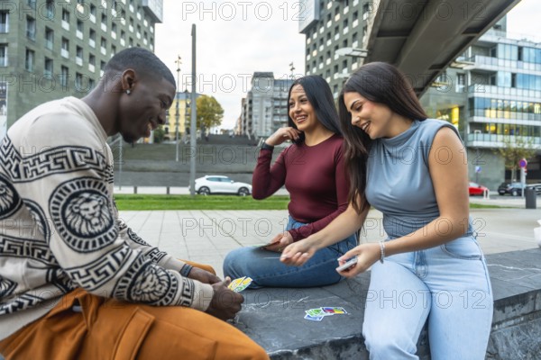 Diverse group of friends sitting outdoors in the city, smiling and laughing while playing cards,