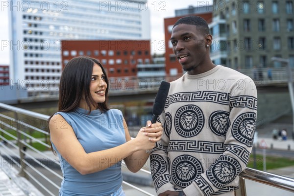 Female journalist interviewing a young black man on a city street, holding a microphone while he
