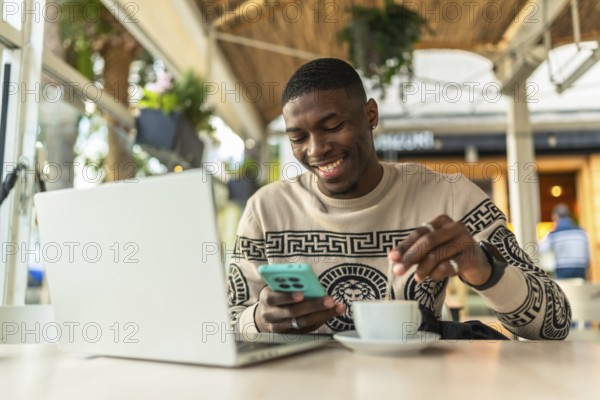 Young black man in a coffee shop using a laptop and smartphone, stirring his coffee while smiling,