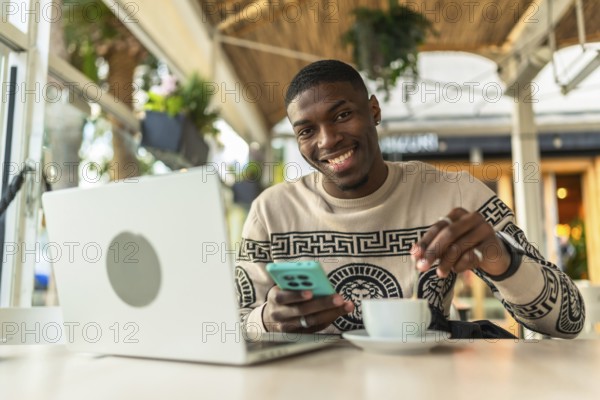 Smiling black man sitting at a cafe with laptop and smartphone, stirring coffee while working