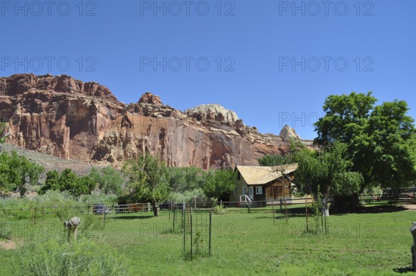 Small wooden house in front of reddish brown rocks, surrounded by green trees and grass, Fruita,