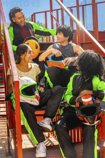 Happy diverse friends group wearing karting suits and helmets, sitting on metal stairs, laughing