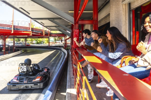 Group of excited diverse friends cheering for a go kart speeding on an indoor multi level track,