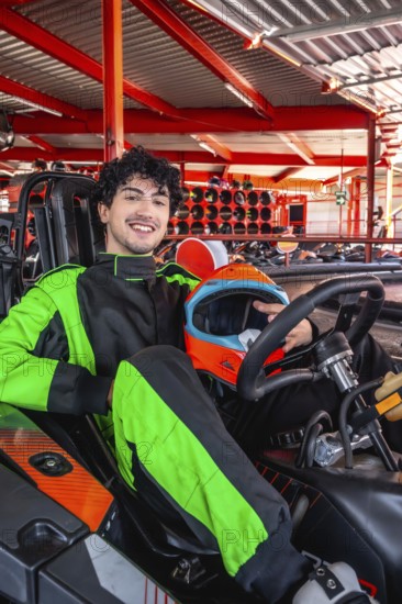 Young man in green and black racing suit smiles holding orange and blue helmet while seated in a go