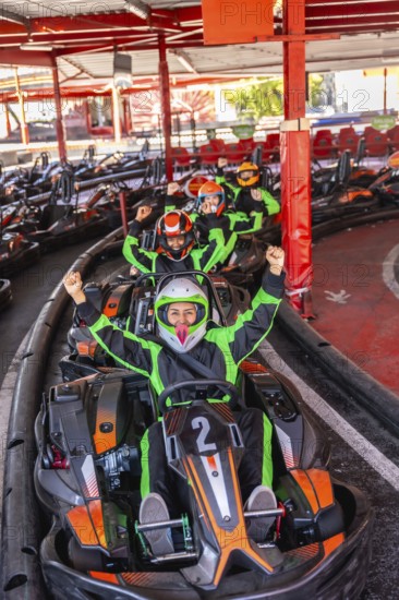Group of excited people wearing helmets and racing suits, sitting in go karts and raising their