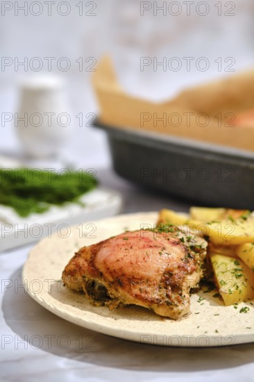 A plate holds a crispy chicken thigh next to golden roasted potatoes. Fresh green herbs are
