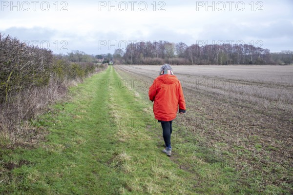 Person wearing bright orange coat walking along grassy path next to hedgerow and winter field,