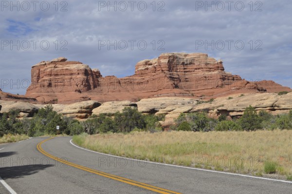 A winding country road runs in front of an impressive red rock formation under cloudy skies,