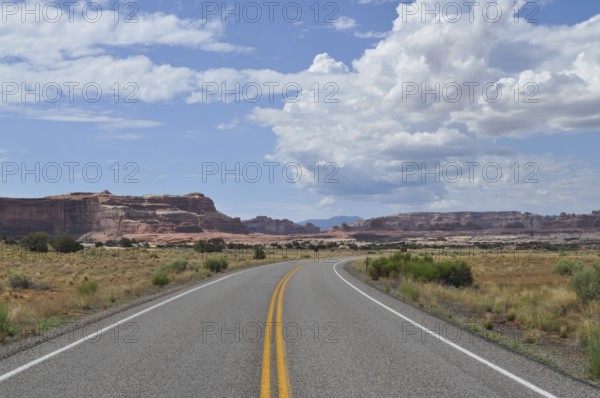 A road leads through vast desert landscape with impressive rock formations and cloudy skies,
