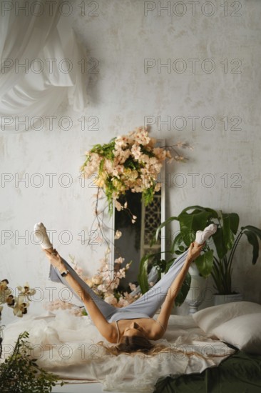 A woman is lying on a bed and stretching her legs in the air. The room has flowers and green plants